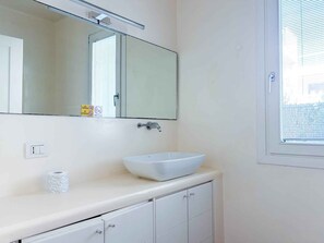 A bathroom with a white sink, a mirror, a window, and a toilet paper dispenser. - Holiday apartment with communal pool (Lido di Jesolo)