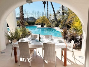 An outdoor dining area with a view of a swimming pool, surrounded by palm trees and a white building. - Holiday apartment with communal pool (Lido di Jesolo)