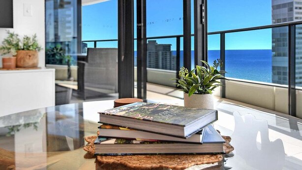A modern room with a glass table, a stack of books, a potted plant, and a view of the ocean and buildings. - Ocean Getaway at Oracle Level 28 (Gold Coast)