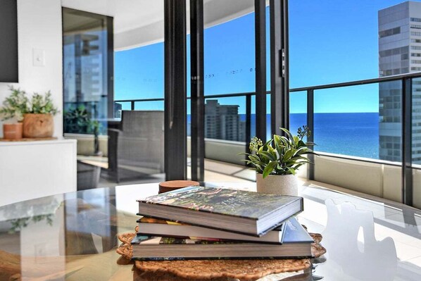 A modern room with a glass table, a stack of books, a potted plant, and a view of the ocean and buildings. - Ocean Getaway at Oracle Level 28 (Gold Coast)