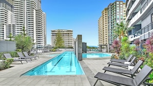 A swimming pool area with lounge chairs, surrounded by tall buildings and greenery. - Beach Breeze Skyhome at Oracle Level 32 (Gold Coast)
