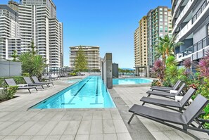 A swimming pool area with lounge chairs, surrounded by tall buildings and greenery. - Beach Breeze Skyhome at Oracle Level 32 (Gold Coast)