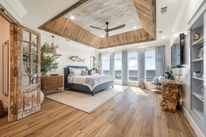 A spacious bedroom with a large bed, wooden ceiling beams, a ceiling fan, and a view of the ocean through large windows. - Bourbon Bay and Whiskey Lullaby Beachfront, Pool, Pickle Ball Court, Fire Pit and Much More (Grand Isle)
