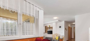 Ein Wohnzimmer mit großem Fenster, einem an der Wand befestigten Fernseher und einer Küchenzeile im Hintergrund.