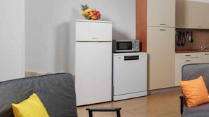 A modern kitchen with white appliances, a microwave, and a small refrigerator. There is a dining area with a wooden table and two grey sofas with yellow cushions. - Holiday flat with balcony and views of greenery (Klimno)
