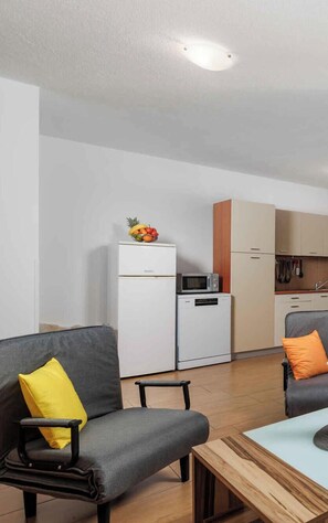 Eine moderne Küche mit weißen Geräten, einer Mikrowelle und einem kleinen Kühlschrank. Es gibt eine Essecke mit einem Holztisch und zwei grauen Sofas mit gelben Kissen.
