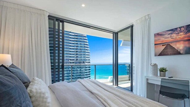 Una camera d'albergo con un letto, una scrivania, una sedia, vista sull'oceano e un edificio sullo sfondo.