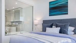 Una camera da letto moderna con un letto grande, un comodino, uno specchio e un bagno visibile attraverso un pannello di vetro.