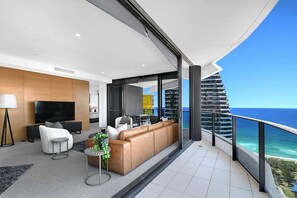 A modern living room with a TV, sofa, and dining area, overlooking a balcony with a view of the ocean and a high-rise building. - Ocean Luxury Skyhome at Oracle Level 36 (Gold Coast)