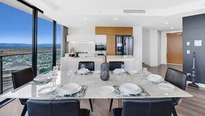 Uma sala de jantar moderna com uma mesa grande posta para seis, uma cozinha ao fundo e vista para a cidade através de grandes janelas.