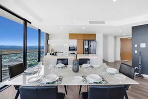 A modern dining area with a large table set for six, a kitchen in the background, and a city view through large windows. - Beach Skyhome at Oracle Level 37 (Gold Coast)