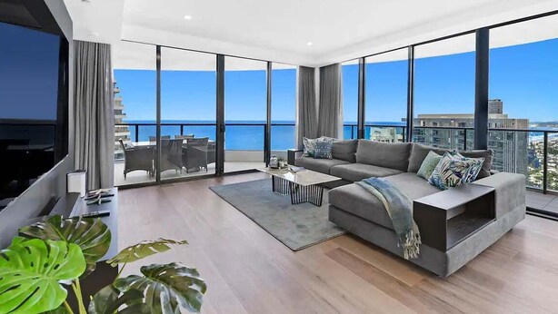 Uma sala de estar moderna com um sofá grande, uma mesa de centro e uma TV. Possui janelas amplas com vista para o mar e edifícios.