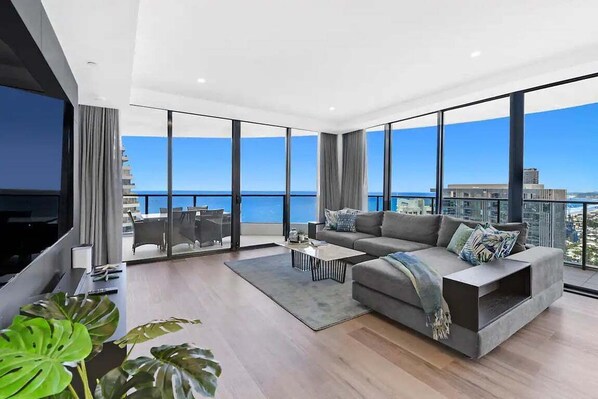 A modern living room with a large sofa, a coffee table, and a TV. It features large windows with a view of the sea and buildings. - Beach Skyhome at Oracle Level 37 (Gold Coast)