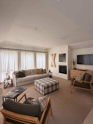 A modern living room with a sofa, ottoman, coffee table, TV, fireplace, and artwork. - Chartres Holiday (Arrowtown)