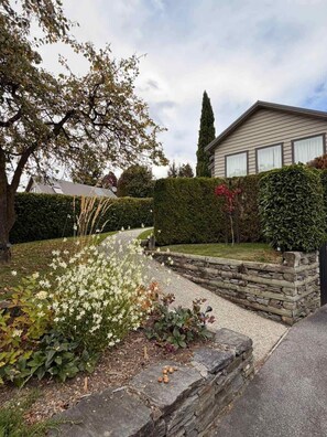 A residential property with a stone retaining wall, a well-maintained garden with white flowers, a neatly trimmed hedge, and a house with a sloped roof and large windows. - Chartres Holiday (Arrowtown)