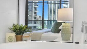 Une chambre d’hôtel avec une grande fenêtre, un lit, une table de chevet avec une lampe, une plante en pot et un petit objet décoratif.