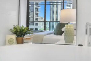 A hotel room with a large window, a bed, a bedside table with a lamp, a potted plant, and a small decorative item.