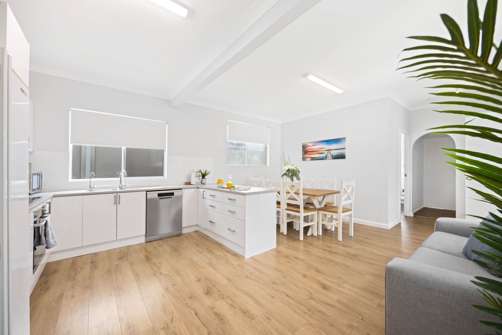 A modern kitchen with white cabinetry, a dining area with a wooden table and chairs, and a grey sofa.