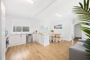 A modern kitchen with white cabinetry, a dining area with a wooden table and chairs, and a grey sofa.