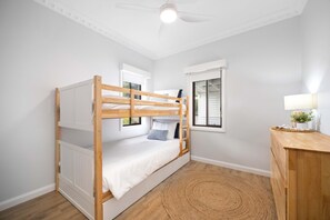A bunk bed with a wooden frame, a white mattress, and a window with blinds. There is a wooden dresser with a lamp and a small plant on top.