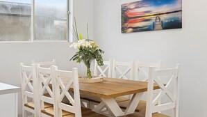 Ein Essbereich mit einem Holztisch und weißen Stühlen, einer Vase mit Blumen und einem Gemälde von einem Sonnenuntergang an der Wand.