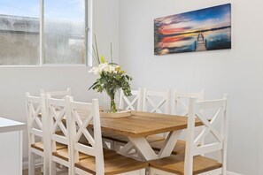 A dining area with a wooden table and white chairs, a vase with flowers, and a sunset painting on the wall.