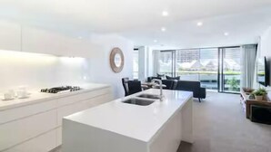 A modern kitchen with a central island, a dining area with a black sofa, and large windows offering a view of the outdoors. - Beach Getaway at Oracle Level 6 (Gold Coast)