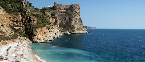 A coastal beach with clear waters, a rocky cliff, and a small boat in the distance.
