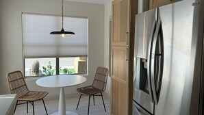 A kitchen with a round table, two wicker chairs, a stainless steel refrigerator, wooden cabinets, and a hanging pendant light. - Norterra Desert Escape: Heated Pool, Walk to Pickleball – Sleeps 5  (Phoenix)