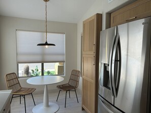 A kitchen with a round table, two wicker chairs, a stainless steel refrigerator, wooden cabinets, and a hanging pendant light. - Norterra Desert Escape: Heated Pool, Walk to Pickleball – Sleeps 5  (Phoenix)