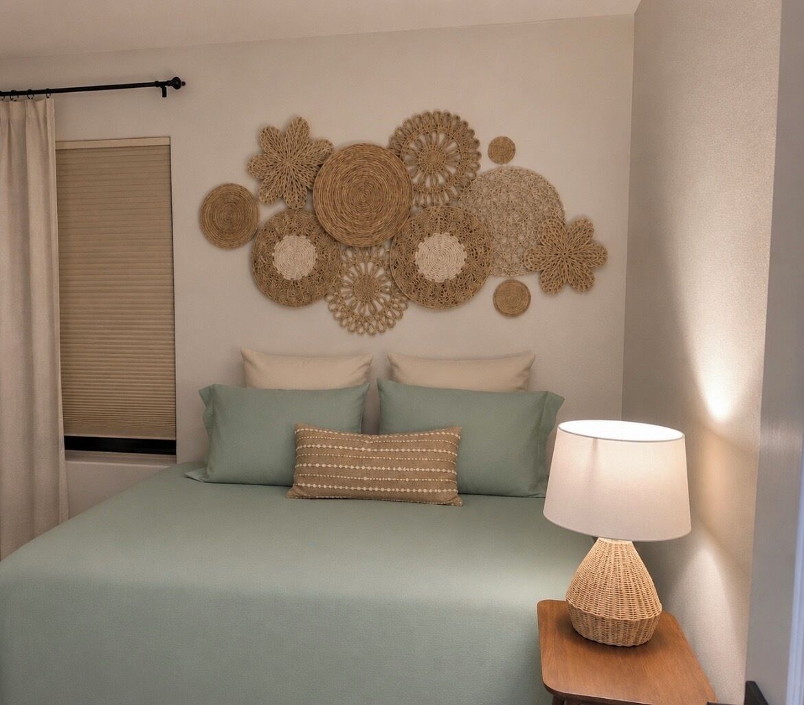 Een slaapkamer met een bed, kussens, een wanddecoratie, een raam met jaloezieën en een bedlamp.