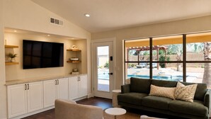 A modern living room with a flat-screen TV, white cabinets, a green sofa, and a view of a swimming pool. - Norterra Desert Escape: Heated Pool, Walk to Pickleball – Sleeps 5  (Phoenix)
