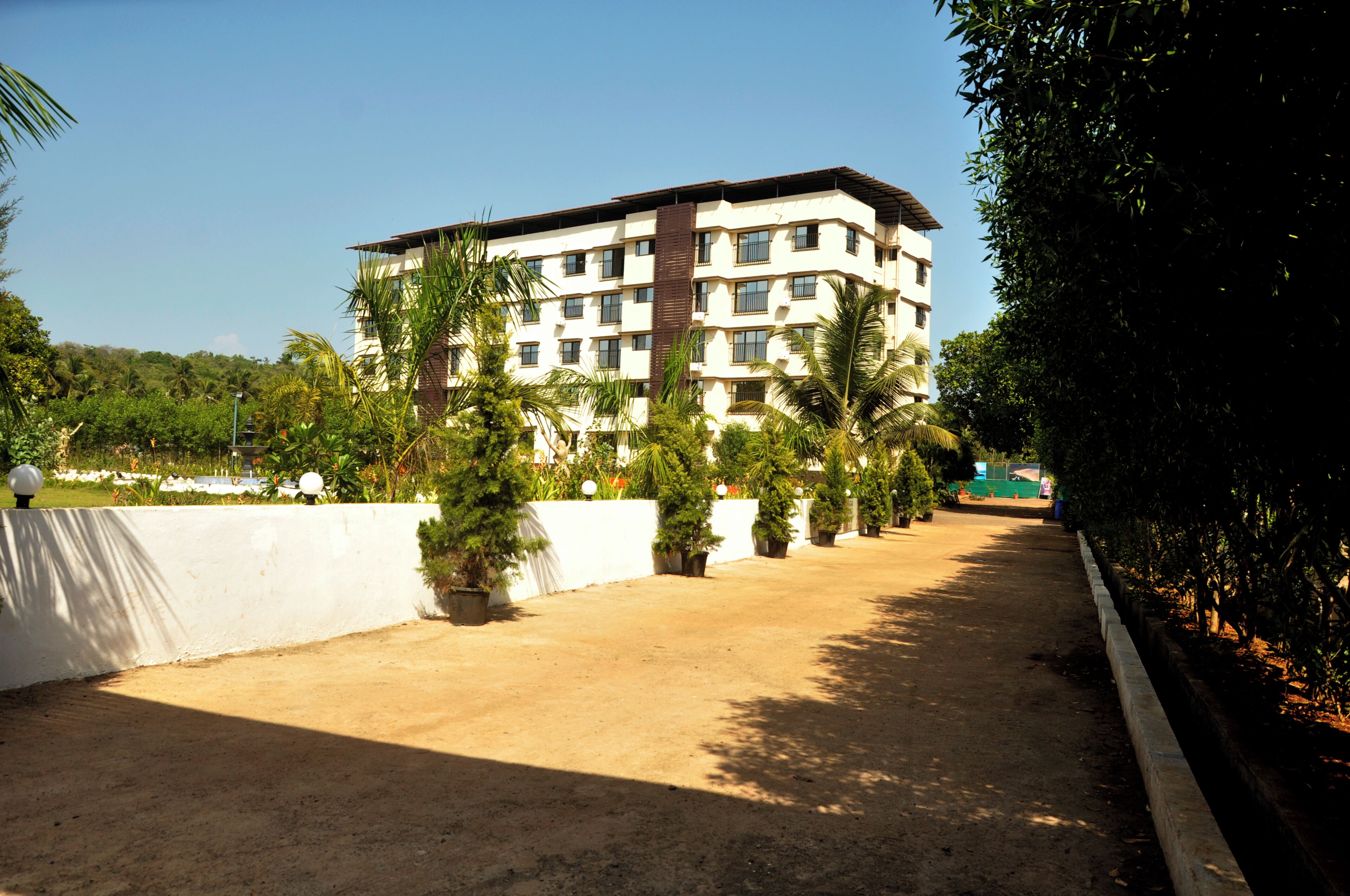 A multi-story building with a flat roof, surrounded by a landscaped area with palm trees and a white wall.