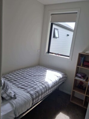 Ein kleines Zimmer mit einem Einzelbett, einem Fenster, einem Bücherregal und einer Steckdose an der Wand.