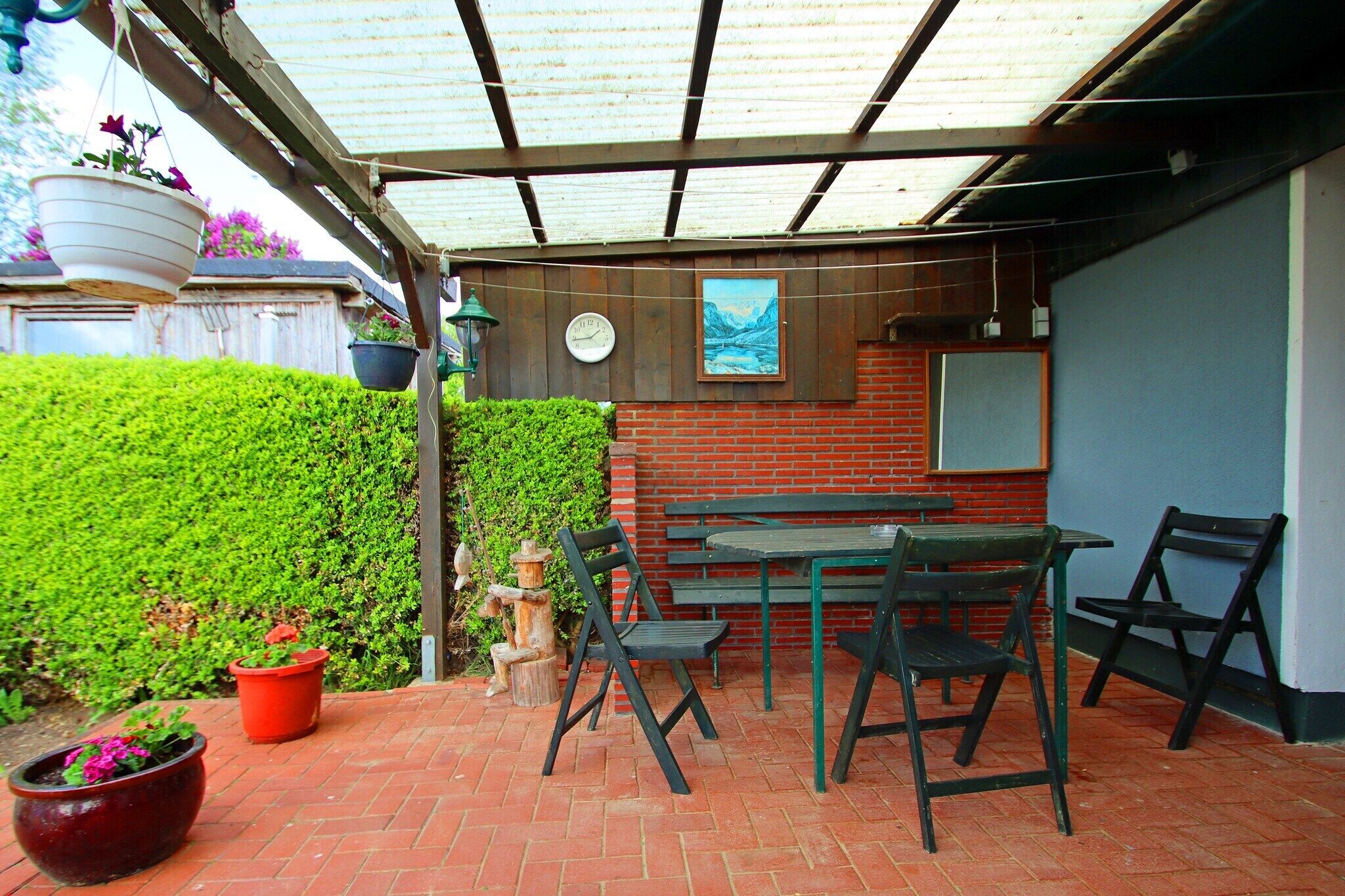 A patio area with a brick floor, green metal chairs, a table, hanging flower pots, a clock, and a framed picture on the wall.