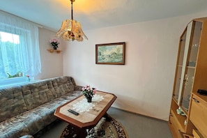 Uma sala de estar com sofá, uma mesa com controle remoto, um lustre, uma moldura com quadro e um armário.