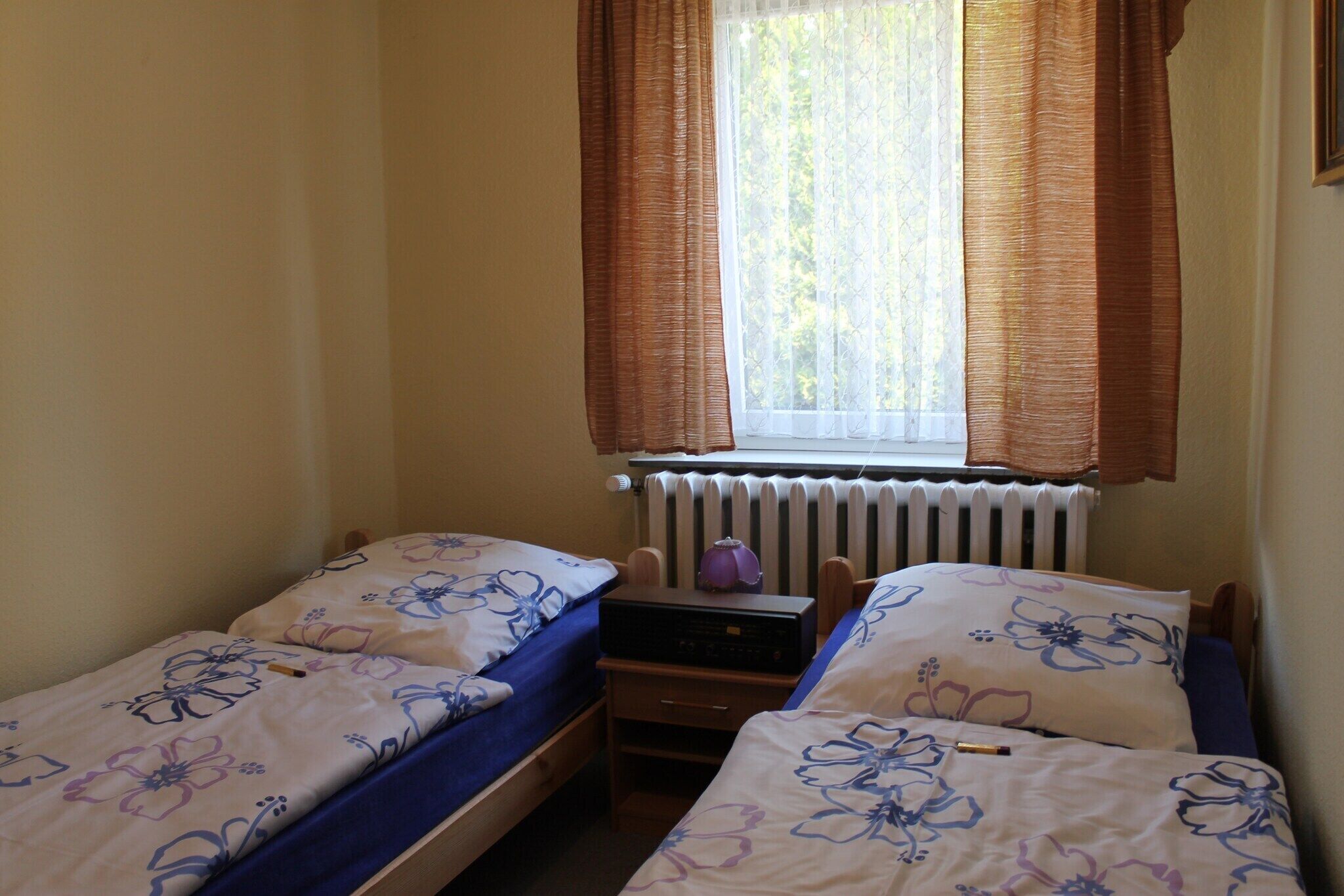 A bedroom with two single beds, a window with curtains, a radiator, and a desk with a lamp.