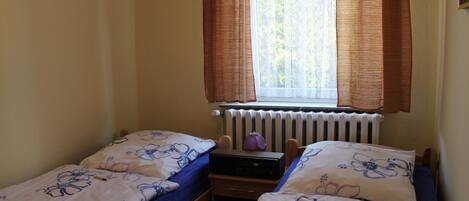 A bedroom with two single beds, a window with curtains, a radiator, and a desk with a lamp.