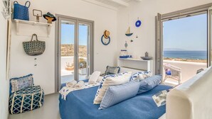 Quarto com cama, poltrona, prateleira com objetos decorativos e vista para o mar.