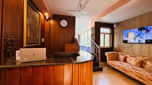 A hotel lobby with a reception desk, a clock, a television displaying multiple rooms, and a seating area with a sofa.