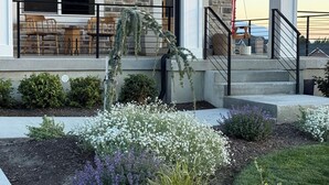 Una casa moderna con fachada de piedra, un porche cubierto con mobiliario exterior y un jardín ajardinado con diversas plantas.