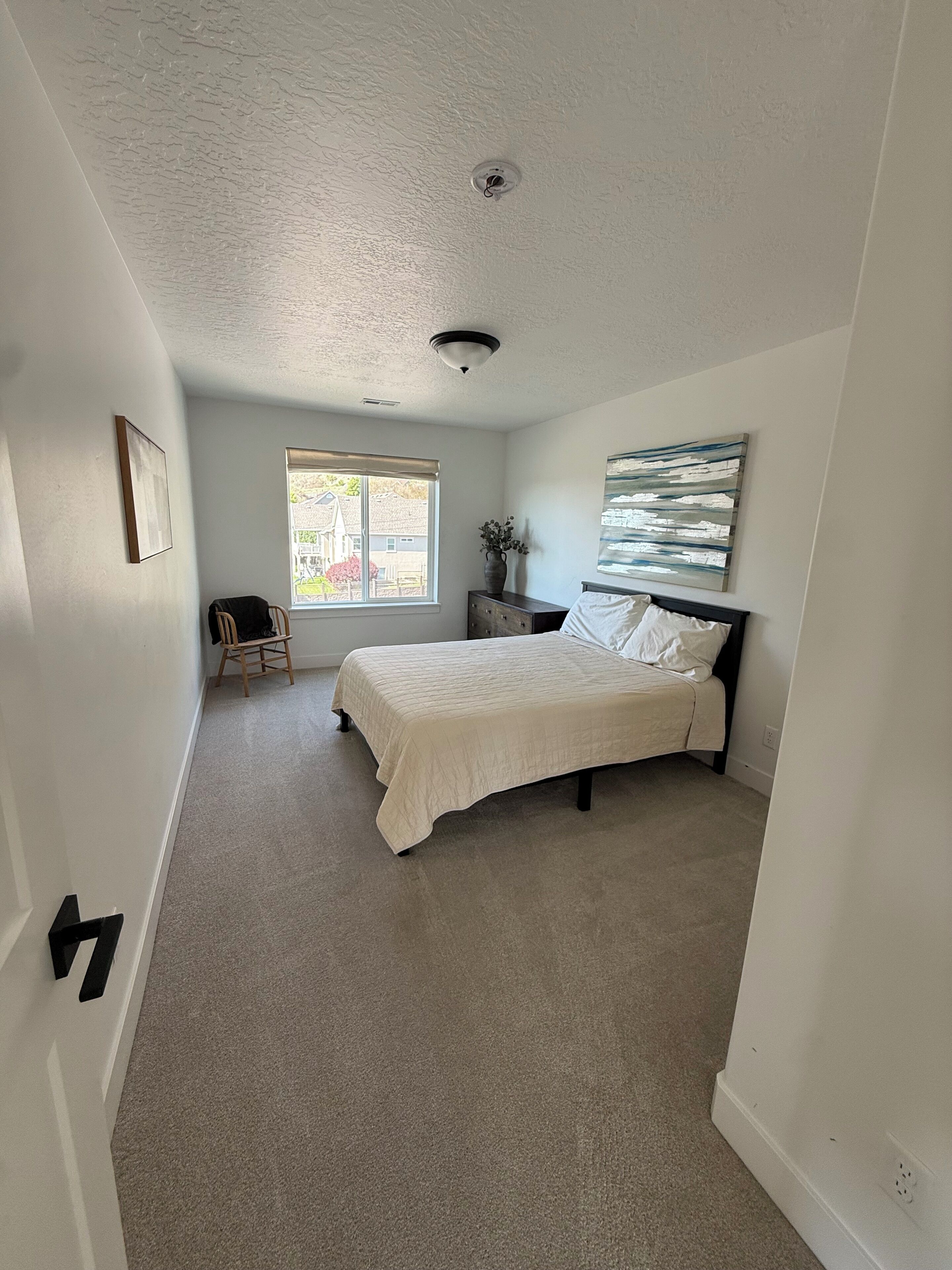A bedroom with a bed, a chair, a window, a painting, and a ceiling light.