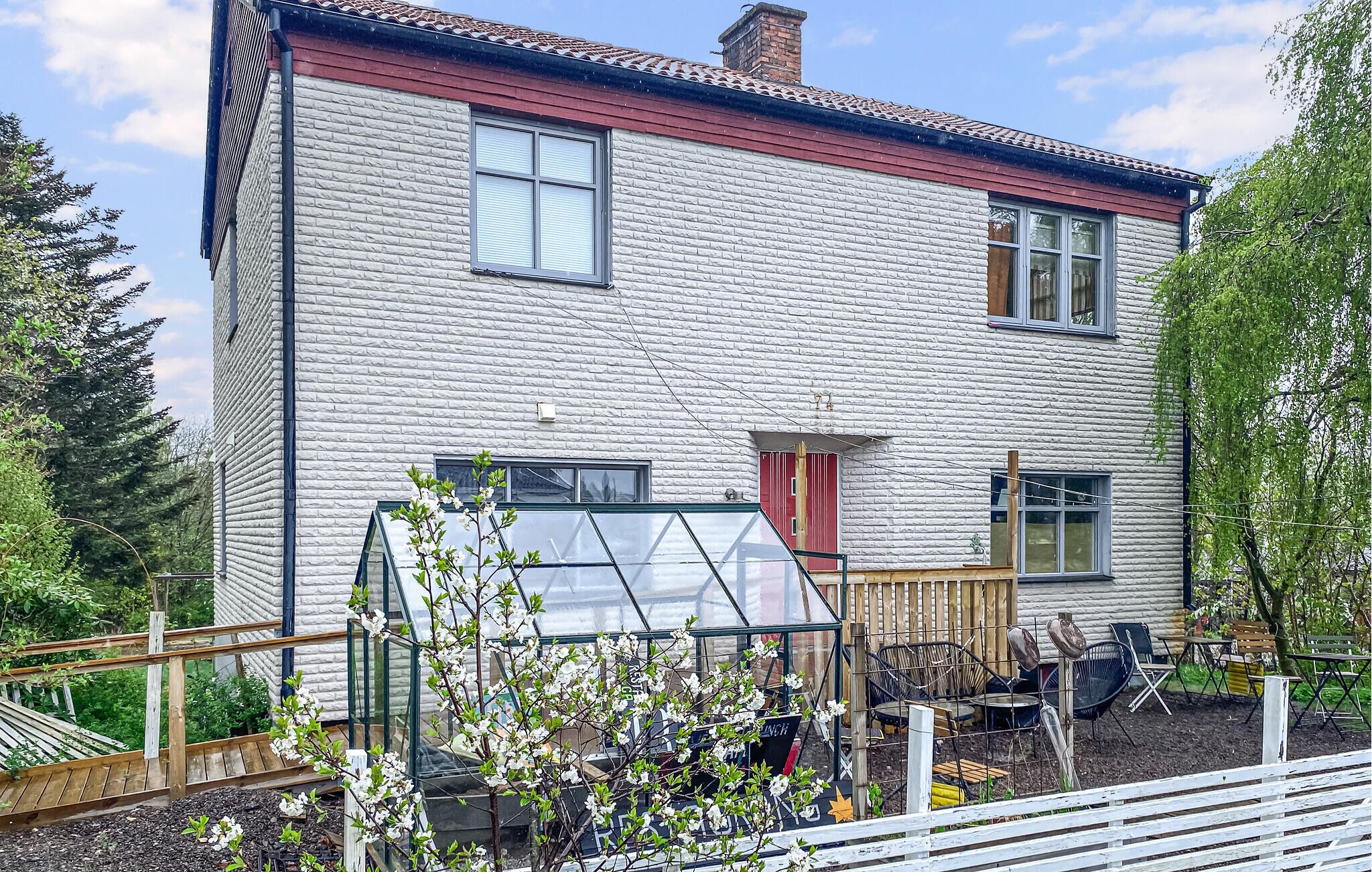 Une maison à deux étages, avec une façade en briques, une porte rouge et un petit jardin agrémenté d’une serre.