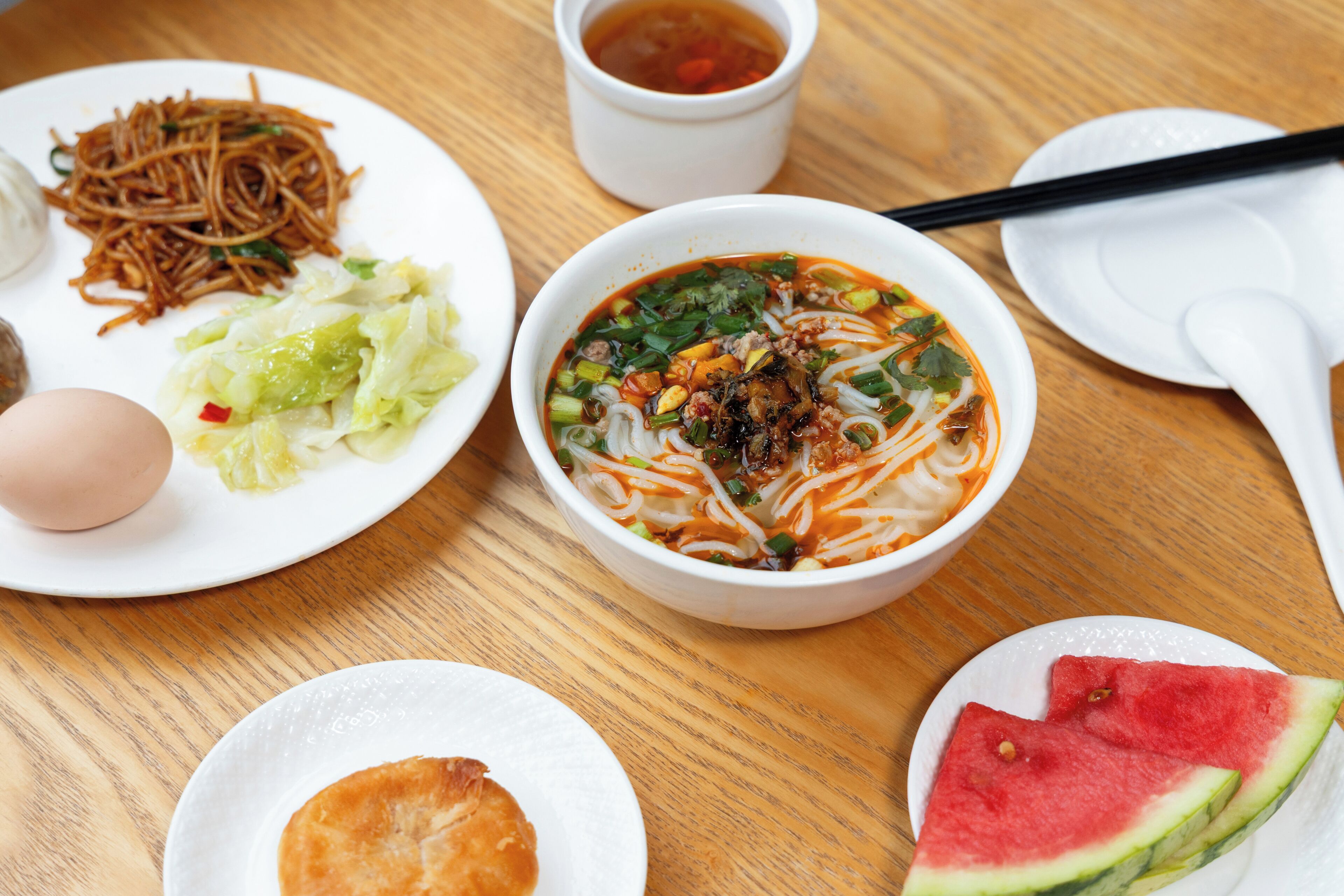 A table with various dishes including noodles, a bowl of soup, a slice of watermelon, and a plate with a boiled egg.