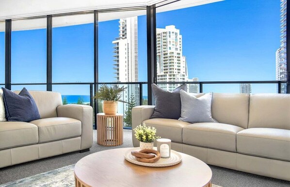 A modern living room with a large window, two sofas, a small round table, and a potted plant. - Ocean Sunrise at Oracle Level 9 (Gold Coast City)