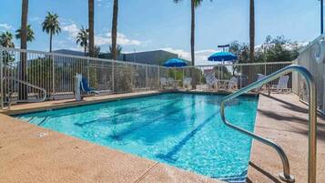 A swimming pool with a metal handrail, surrounded by a white fence, palm trees, and blue umbrellas.