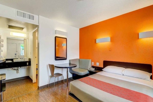 Habitación de hotel con cama, escritorio, silla, mesita pequeña, espejo y mesita de noche.