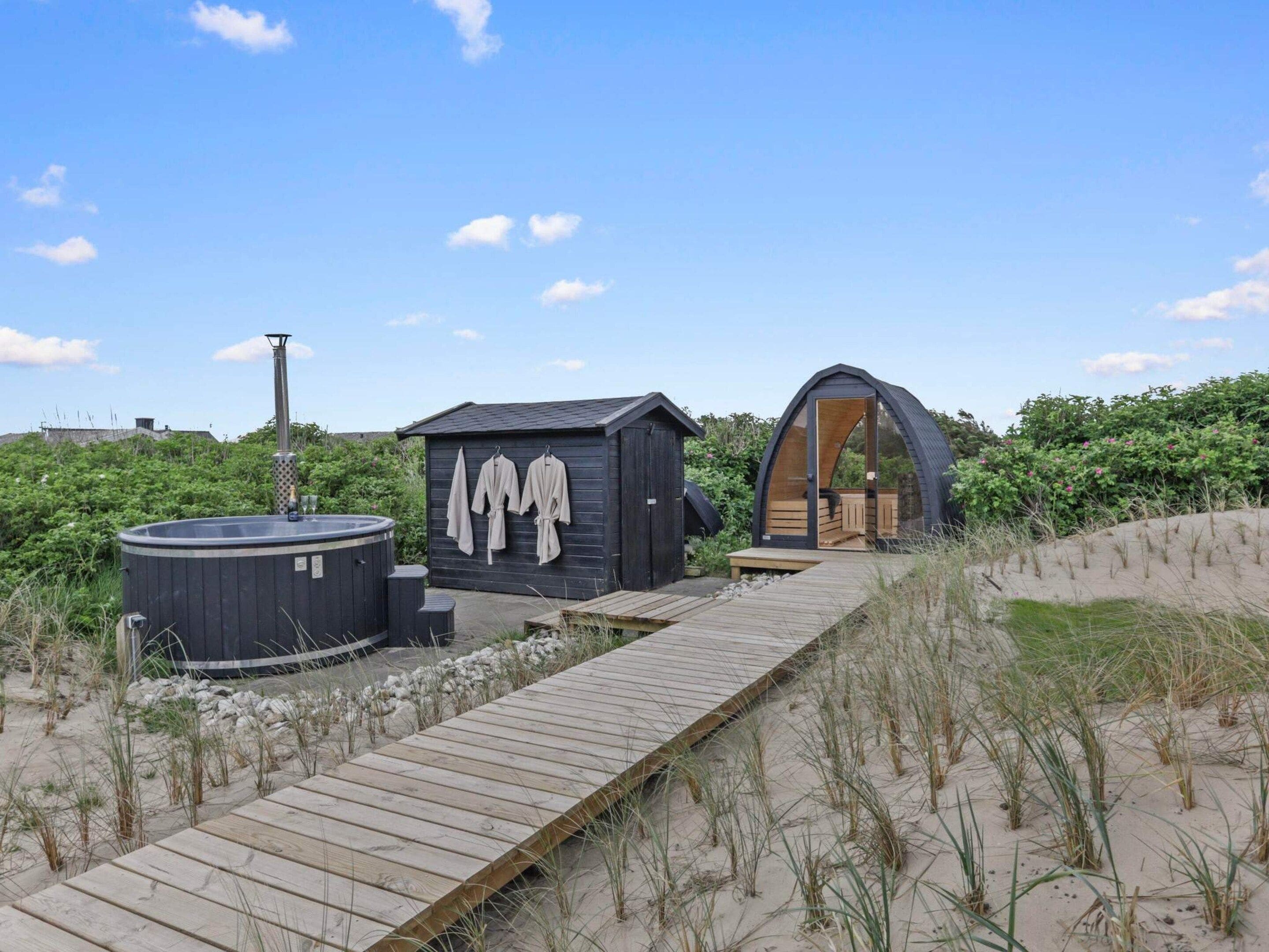 A wooden pathway leading to a beachside structure with a hot tub and a small building with a curved entrance.