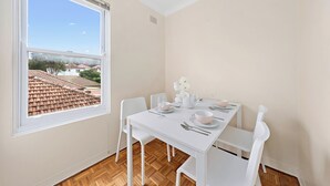 Een eetruimte met een tafel voor vier personen, witte stoelen, een raam met uitzicht op een woonwijk en een ventilatie rooster in het plafond.