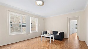 Ruime kamer met een visgraatparketvloer, een witte salontafel, een zwarte bank en een raam met uitzicht op een bakstenen gebouw.